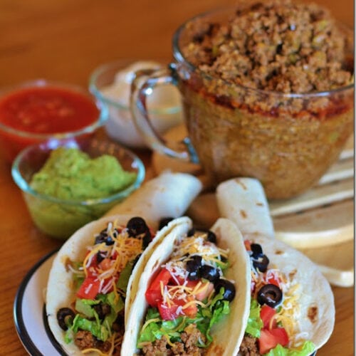 Three Full tacos on a round plate sitting in fron of a glass bowl of taco filling and other fixings