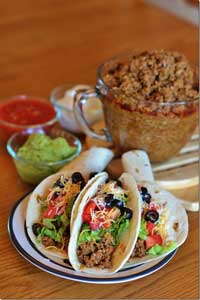 Round plate with three stuffed tacos and a large glass bowl of taco filling in the background