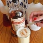 Pumpkin Spice seasoning being added to Hot Buttered rum beverage in a clear glass sitting on a table in front of two bottles of rum