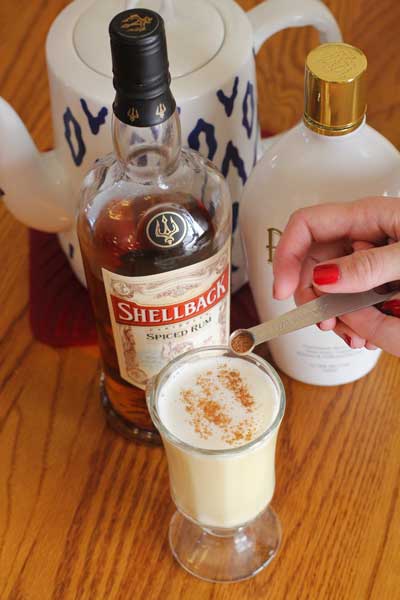 Pumpkin Spice seasoning being added to Hot Buttered rum beverage in a clear glass sitting on a table in front of two bottles of rum