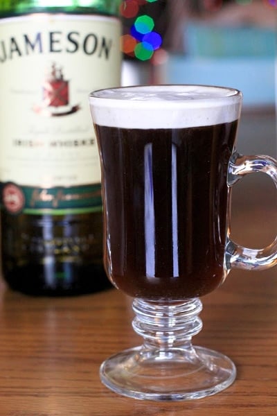 Authentic Irish Coffee in clear glass set on a wood table with a bottle of Irish whiskey in the background