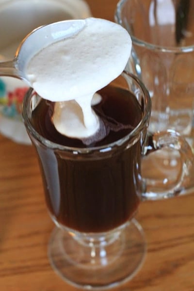 Spoon full of homemade whipped cream being added to Authentic Irish Coffee in clear glass 