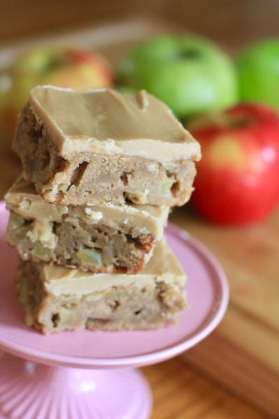 three Maple Apple Cake Bars stacked on a pie plate  in front of apples on a wood table