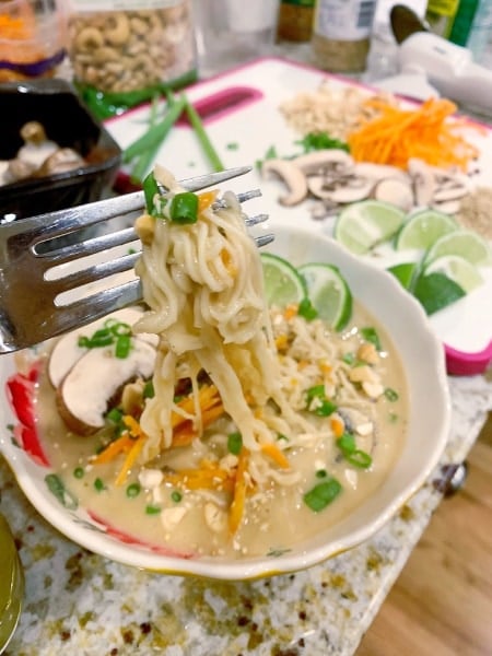 fork full of Spicy Thai Vegetable Ramen above a bowl with a cutting board full of ingredients in the background