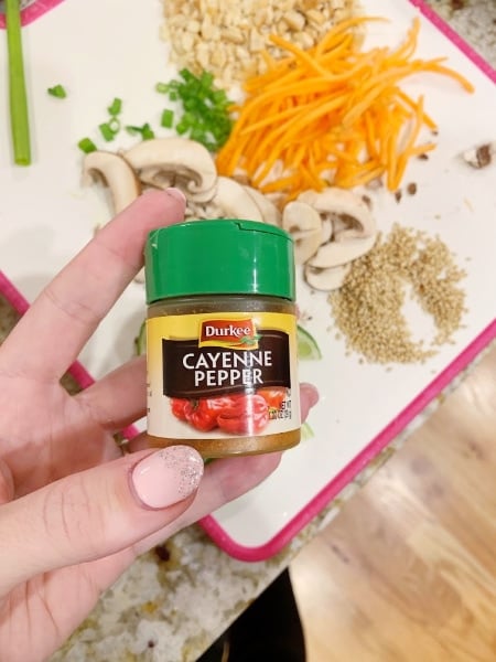 hand holding a container of Cayenne Pepper  above a cutting board full of chopped ingredients for Spicy Thai Vegetable Ramen 