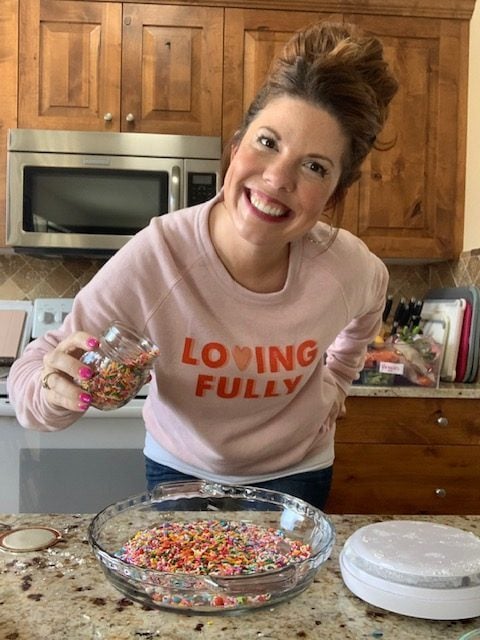 Lexi is holding a jar of sprinkles and wearing a pink sweatshirt, smiling at the camera