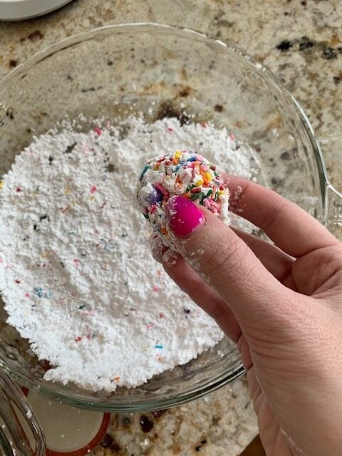 Lexi's hand holding a colorful cookie dough ball and a plate of powdered sugar.