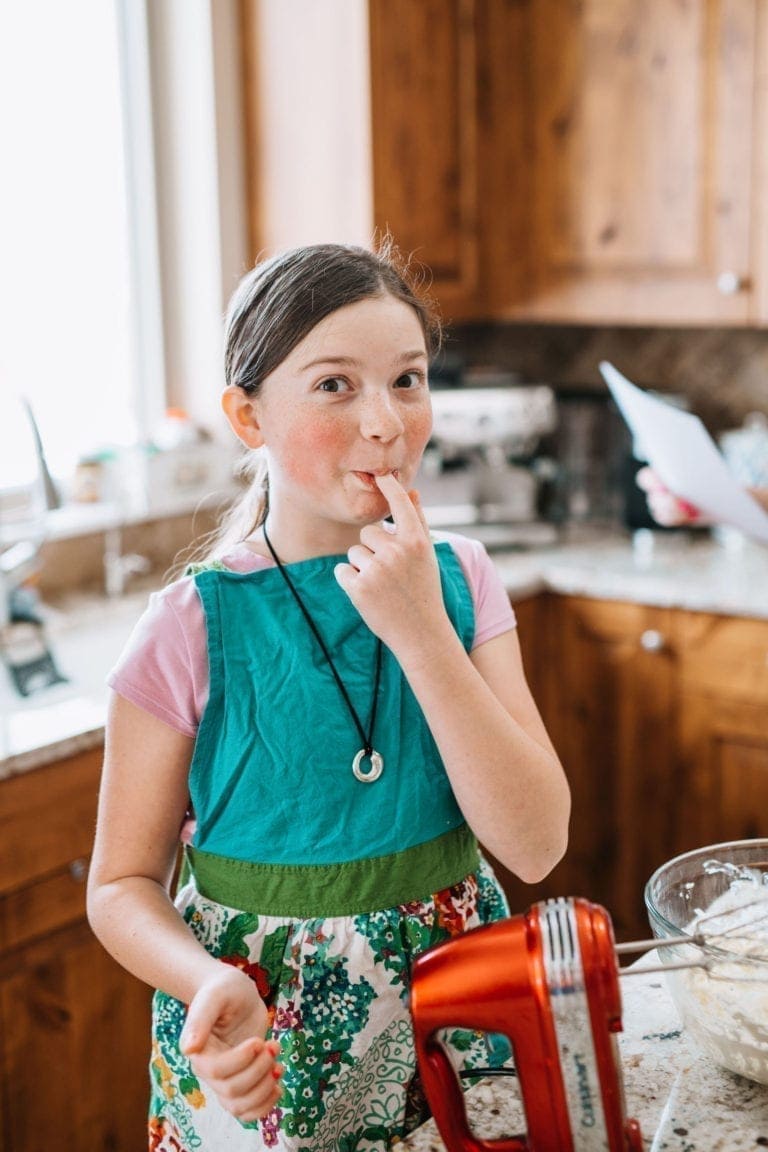 Young girl standing in kitchen with an apron smiling at camera as she takes a tast of the cake batter she is making