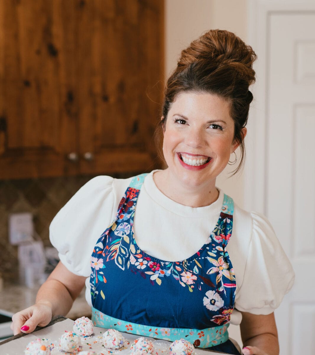 Lexi is wearing an apron, holding a tray of cookies and smiling at the camera.