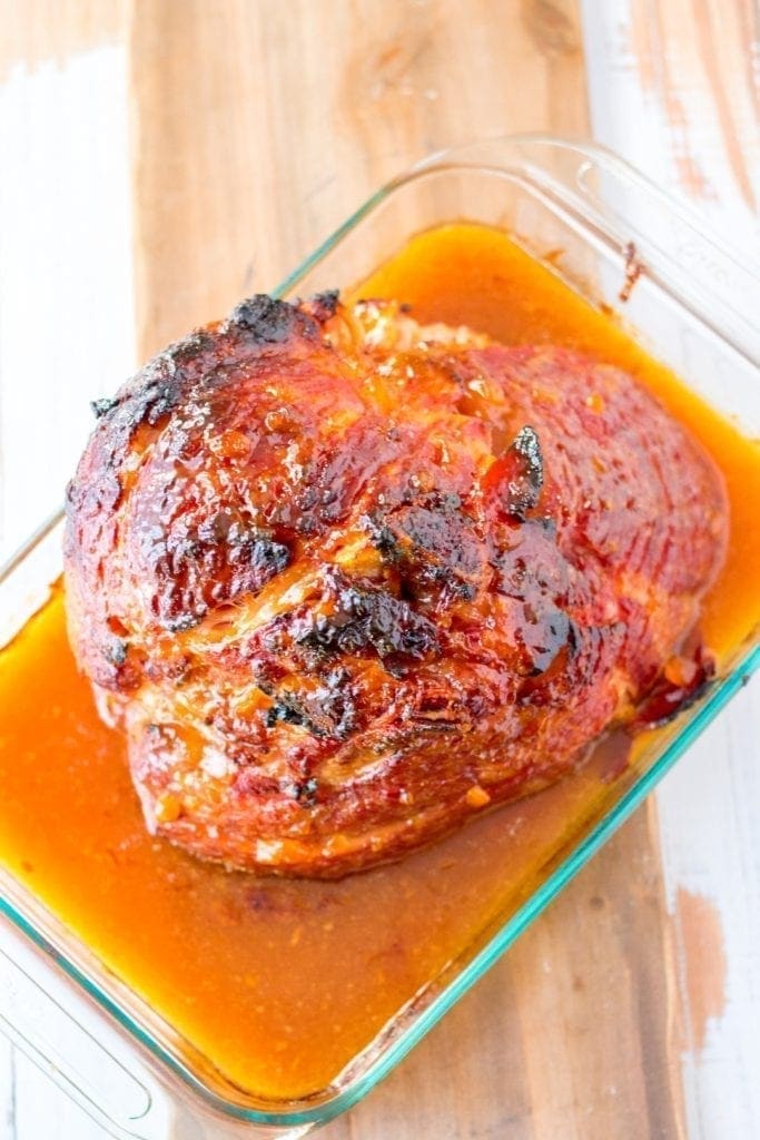 Apricot dijon Glazed Ham bathing in its juices cooling on a white, wood table. 