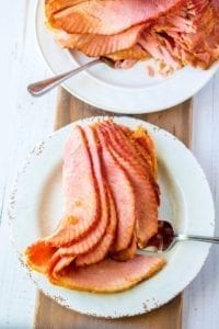 Apricot dijon Glazed Ham carved sitting on a white plat with fork on cutting board