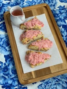 Strawberries and Cream scones text overlay with an image of four scones covered in red frosting sitting on a wood tray with blue table cloth