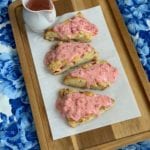 Strawberries and Cream scones text overlay with an image of four scones covered in red frosting sitting on a wood tray with blue table cloth
