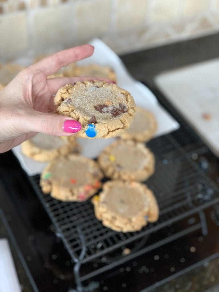 Fresh cookies held in hand with other cookies on cooling rack. Cookies dotted with colorful M&ms and sprinkled with sugar.