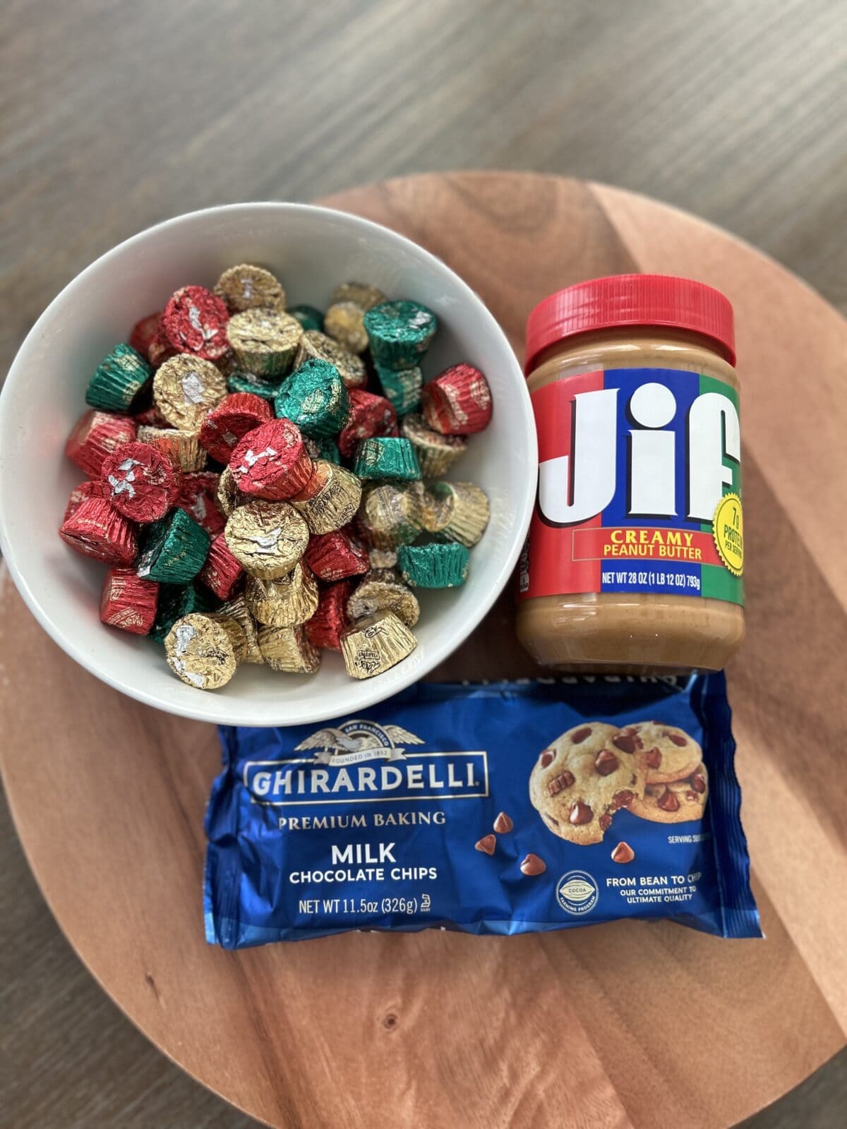 Jar of peanut butter, package of milk chocolate chips and a bowl full of peanut butter cups in a white bowl sitting on a wood tray