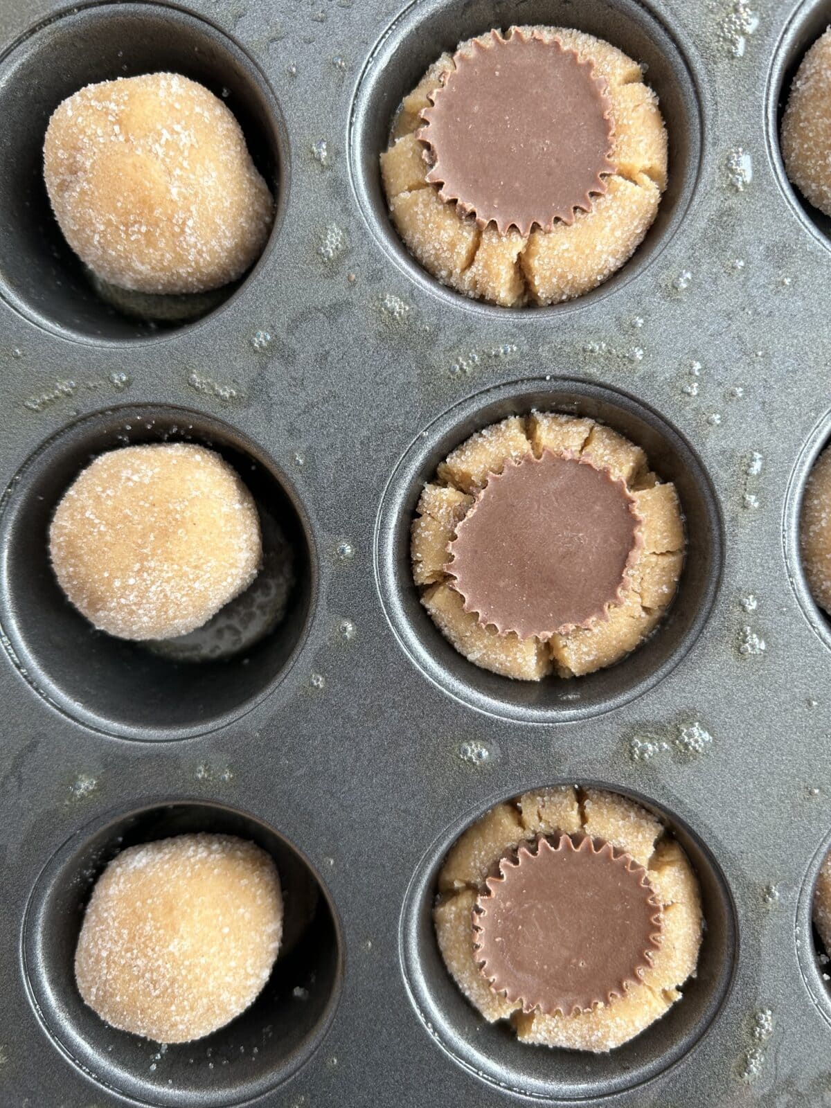 Close up of a mini muffin tray filled with peanut butter cookies. One with just dough and one with the peanut butter cup pressed inside.