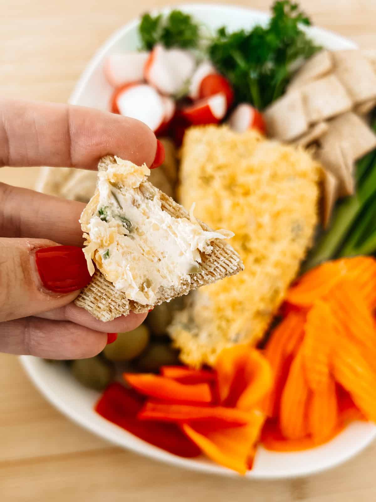 cheese spread on a cracker and a cheese ball on a tray surrounded by veggies and crackers.