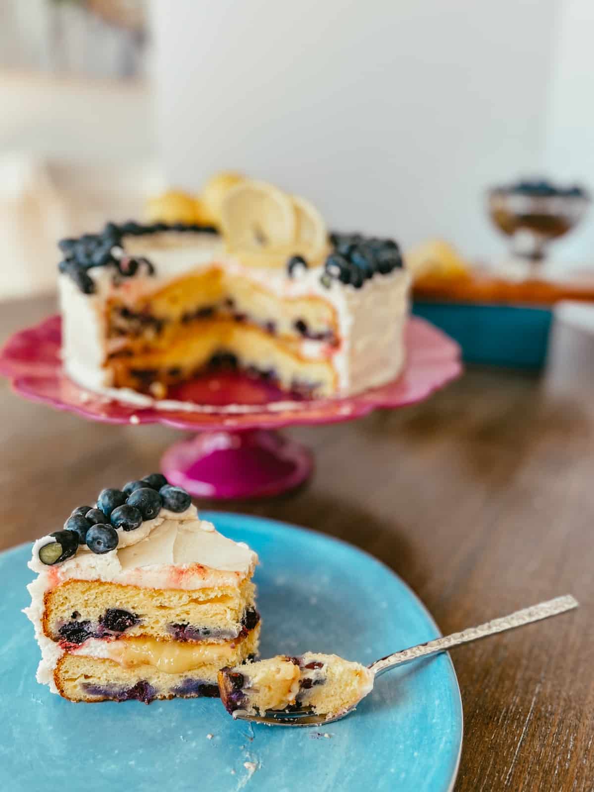 Slice of layered cake with blueberries on a plate and a cake on a pink cake stand