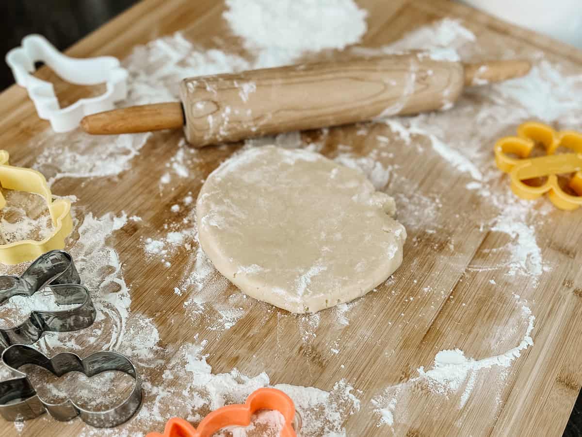 Sugar cookie dough on a wood board with a rolling pin, flour, and cookie cutters.