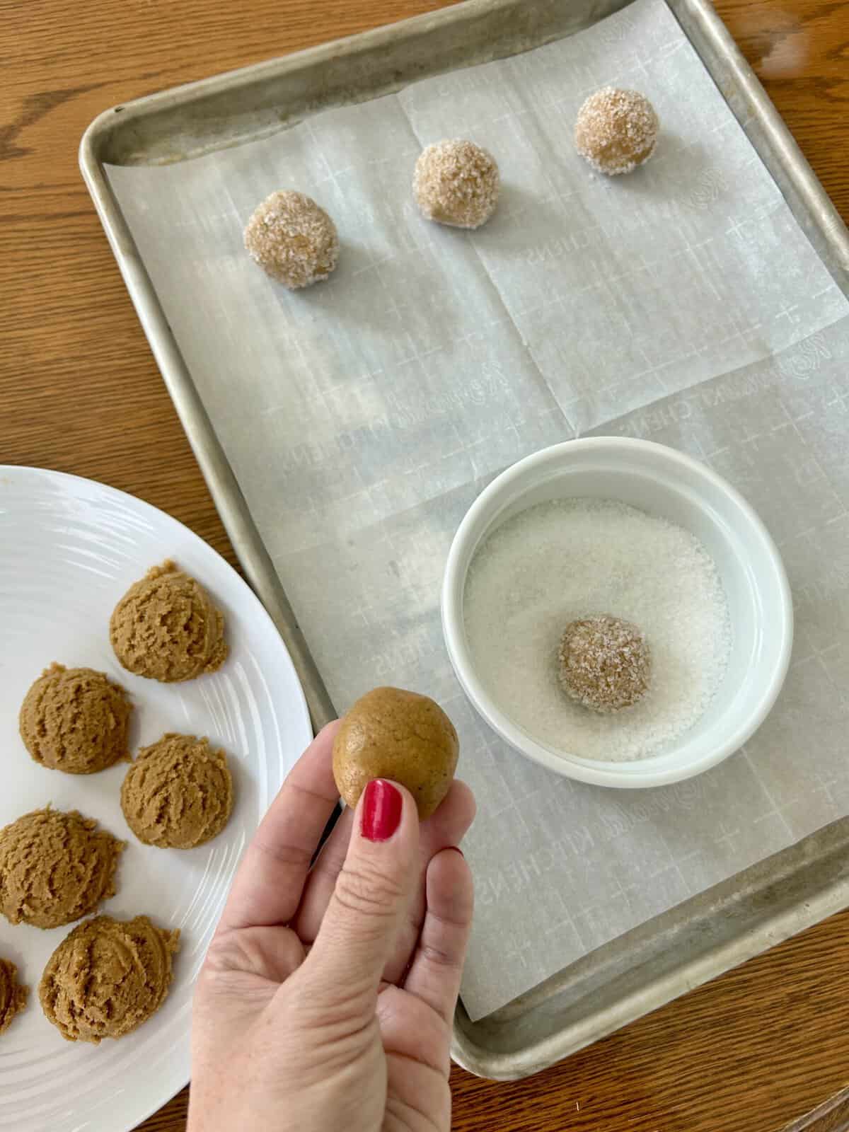 Cookie dough balls on a plate, one in a bowl of sugar and 3 on a baking sheet